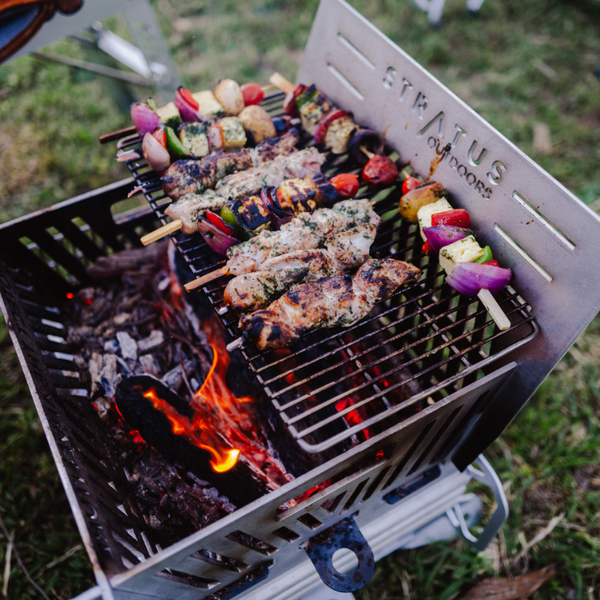 Standard SS grill shown with skewers of chicken, vegetables, and onions cooking over flames and embers on a grassy outdoor surface. The Firepit has 'STRAUS OUTDOORS' laser cut on the back panel.