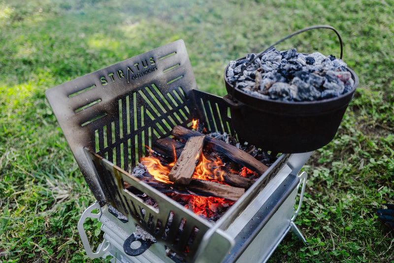 Stratus Firepit Oven set up on grassy area, with visible flames and coals, with a cast iron camp oven placed on top for cooking