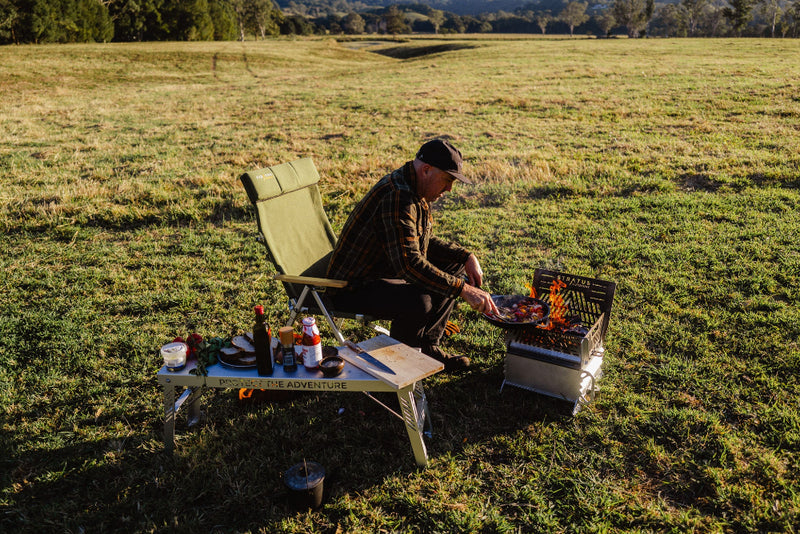 Firepit Oven set up in green field campsite with man cooking vegetables in a cast iron pan over the flames