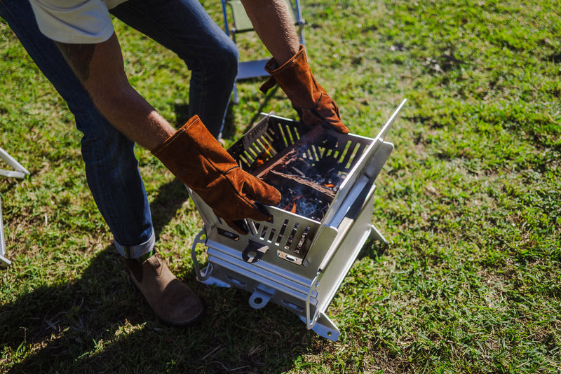A man outdoors on grass wearing brown leather gloves and lifting the Firepit Oven from the side handles with visible embers and fire burning in the firepit.