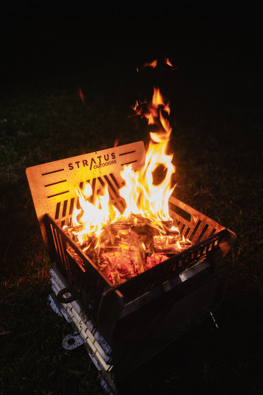 A Stratus Outdoors portable fire pit oven, shown lit with bright orange flames on a dark evening outdoor background.