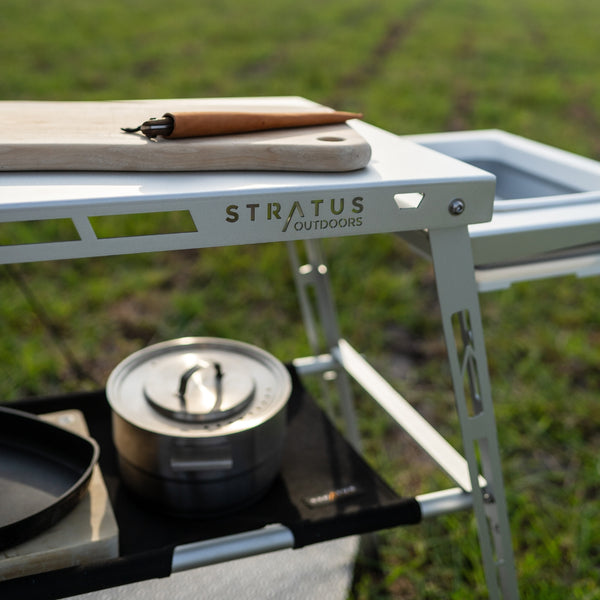 Diagonal angle showing black waxed canvas hammock shelf hanging beneath table, fully loaded with camping gear