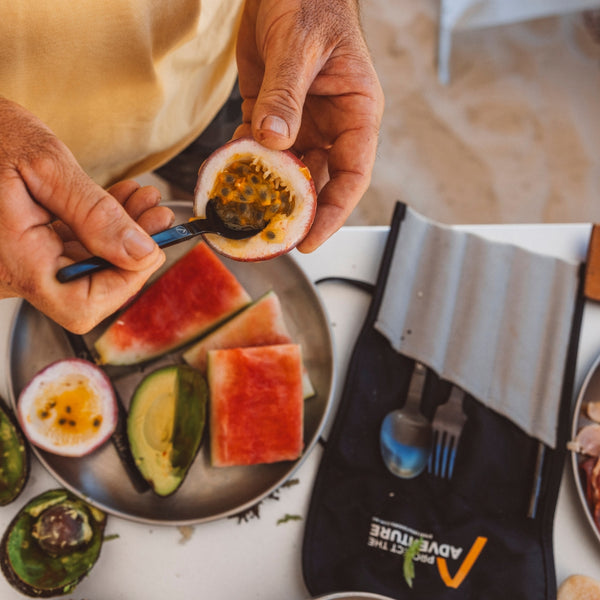 Person holding a halved passion fruit over a plate of sliced watermelon and avocado on a table.