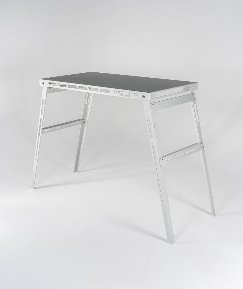 Side-angled view of the Big D camp table showing its 900 mm high brushed aluminium surface and folded stainless steel legs.