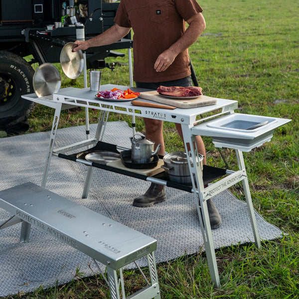 Stratus Outdoors Big D Table in use as a camp kitchen station with food ingredients and cookware arranged on its brushed aluminium surface.