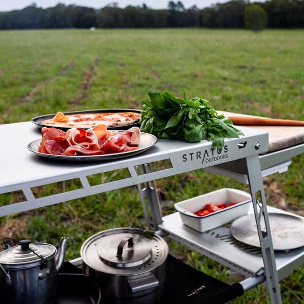 Outdoor cooking scene showing the Stratus Outdoors Big D Table used as a prep bench with food and kitchen tools on surface.