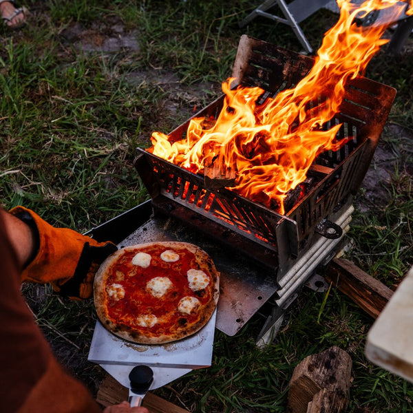 Stratus Outdoors Firepit Oven cooking session: wood fire below, pizza sliding out from oven tray.