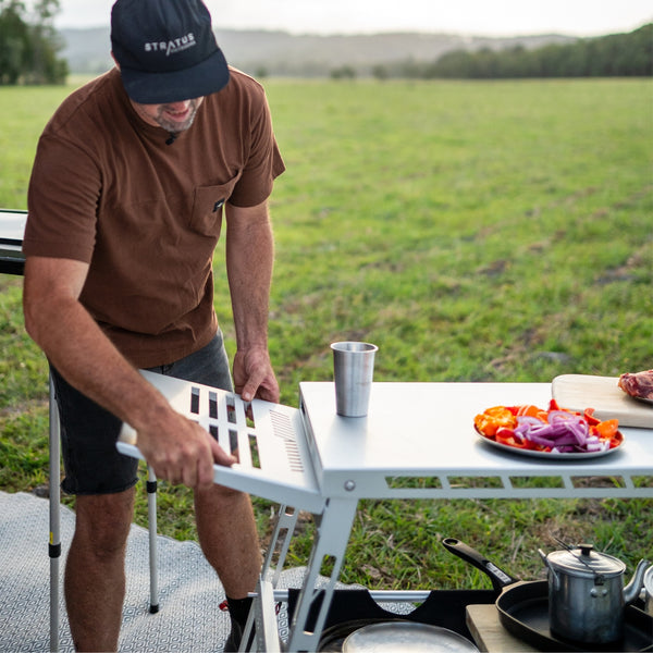 Outdoor kitchen scene: Stratus Outdoors Sink Rack mounted on camp table, featuring chopped vegetables, cookware and water drain board.