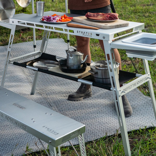 Outdoor scene of Under Table Hammock under camp table, black canvas shelf holding food prep tools.