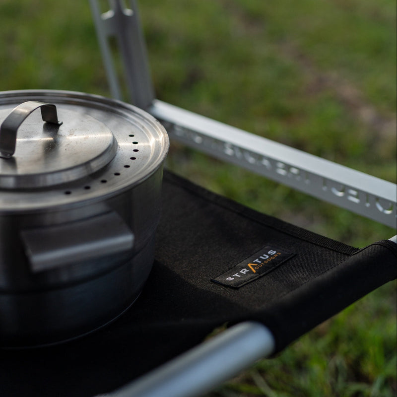 Under Table Hammock in use beneath a camp table, black waxed canvas holding cookware, supported by white alloy poles.